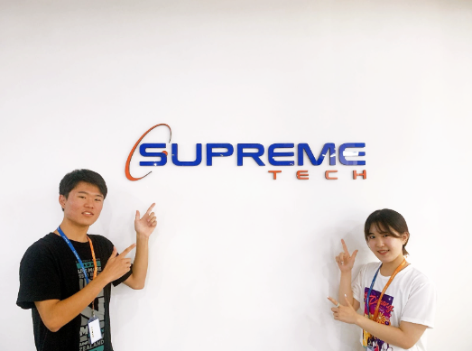SupremeTechのロゴの前に立つインターン生のMさんとSさん